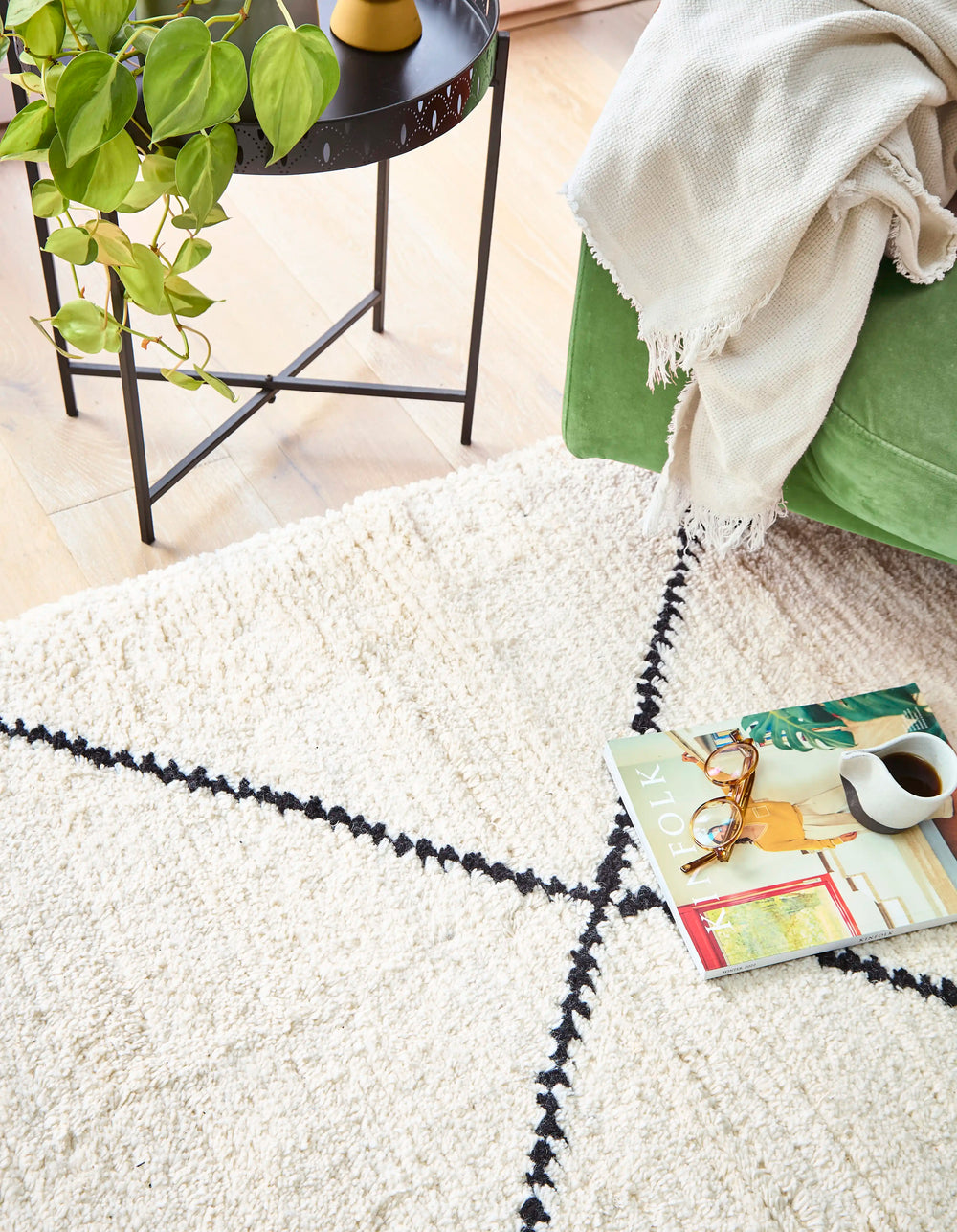 A cream wool berber rug with black diamond pattern in a sunlit room, with a green armchair, book and coffee on top next to a table and plant