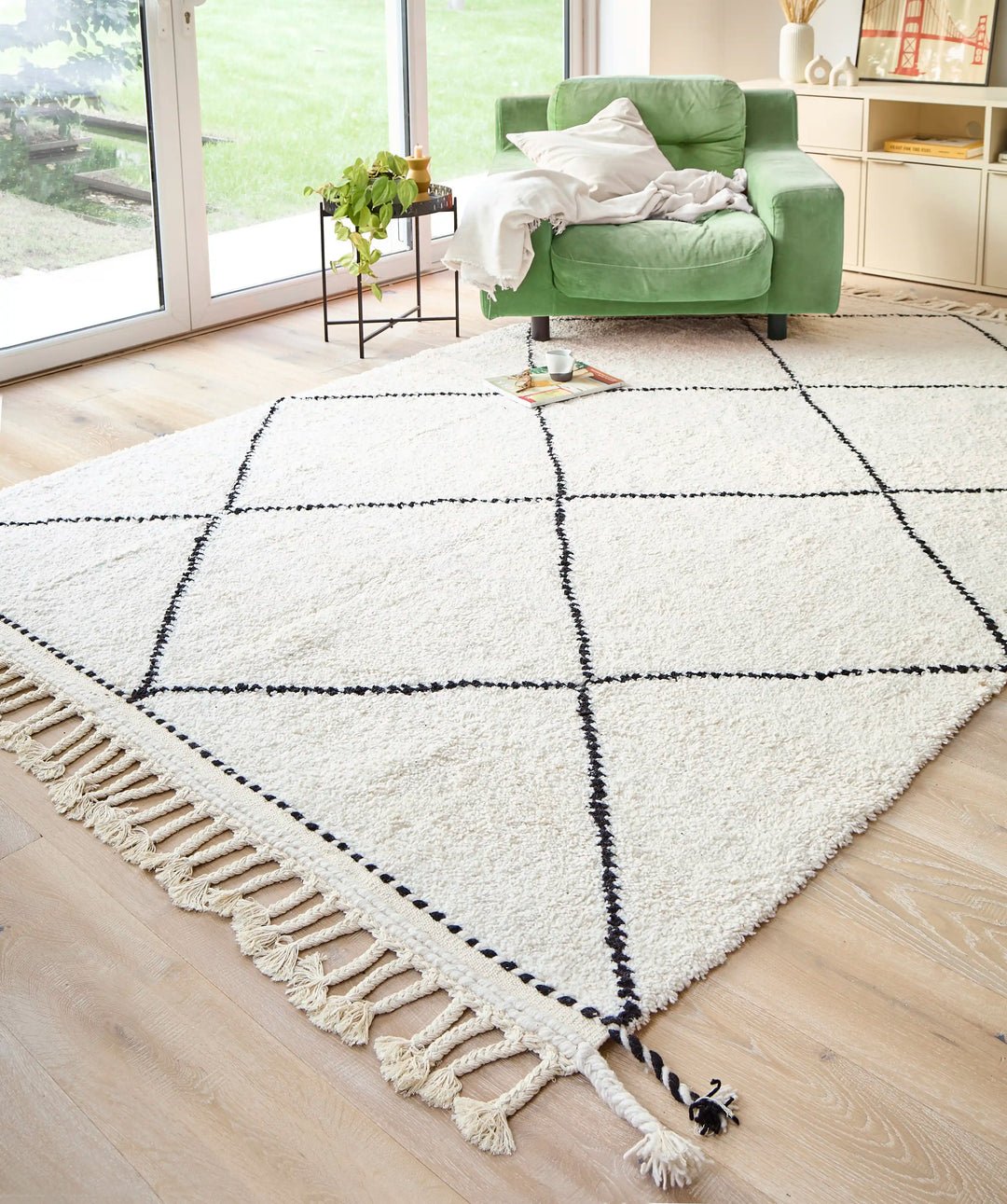 A cream wool berber rug with black diamond pattern and plaited tassels in a sunlit room, with a green armchair on top