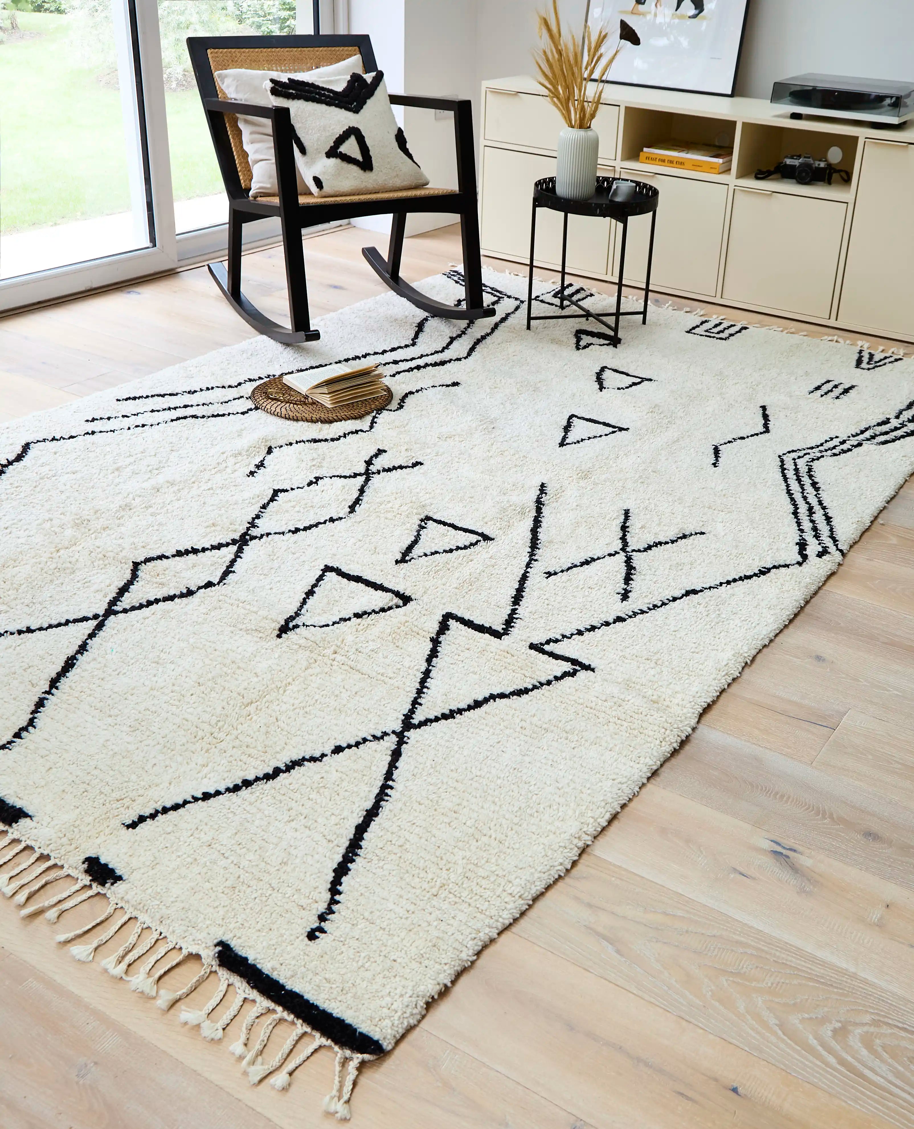 A cream wool berber rug with black abstract shapes on a wooden floor in a living room, with a black rocking chair on top