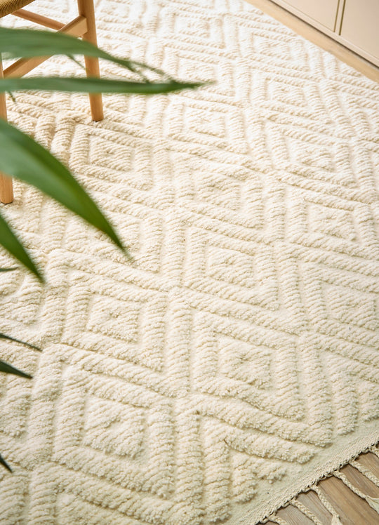 A natural wool cream berber rug with a diamond design on a wooden floor next to a plant.