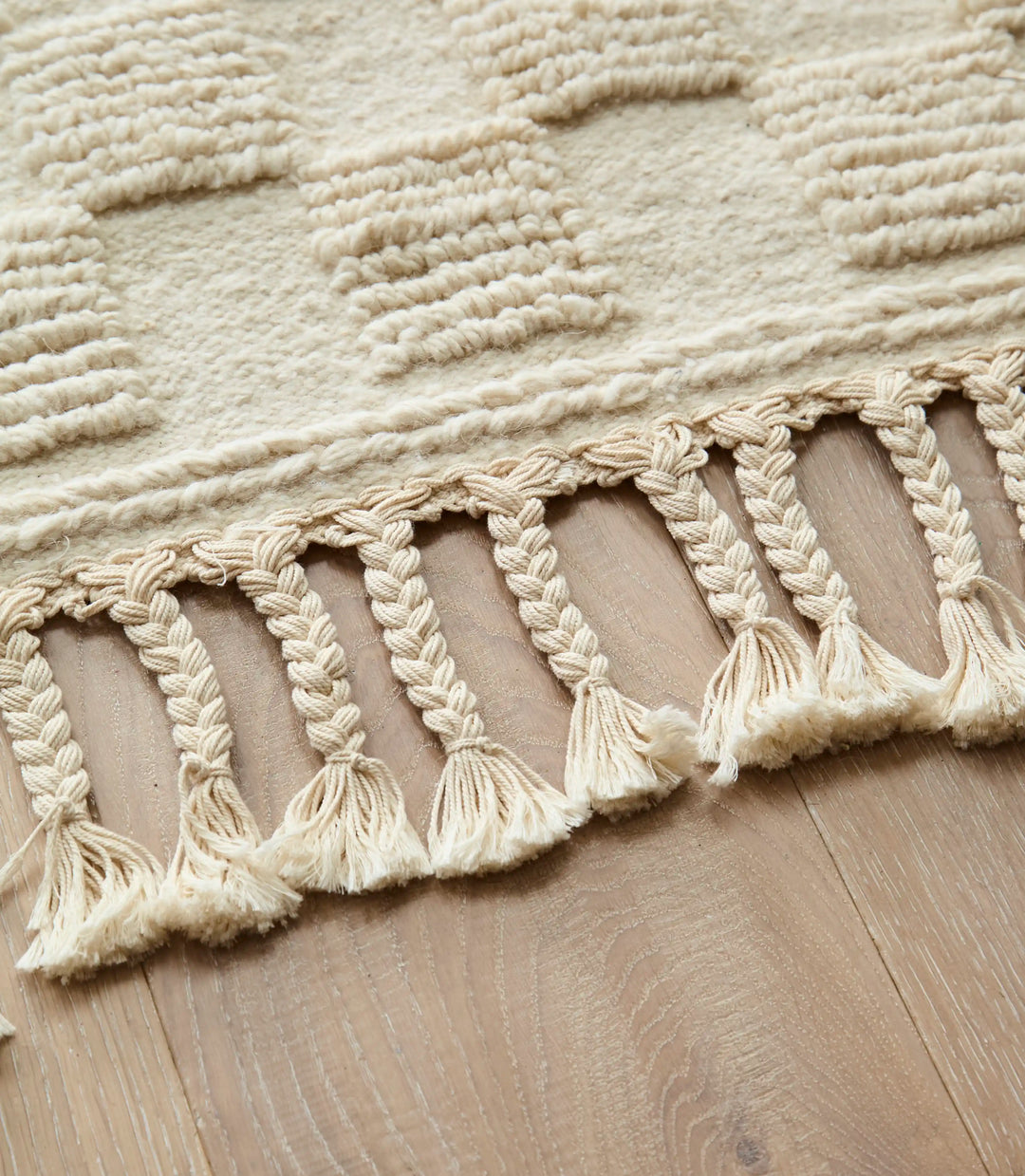 ​Handwoven cream Berber rug with square geometric design, featuring close up on plaited tassels set on a wooden floor.