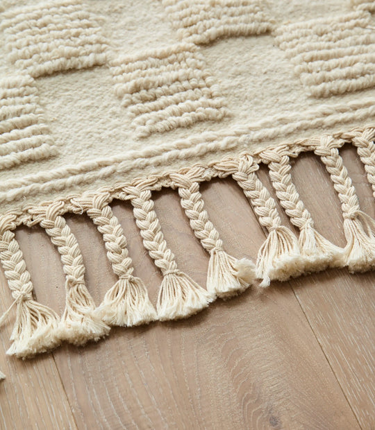 ​Handwoven cream Berber rug with square geometric design, featuring close up on plaited tassels set on a wooden floor.