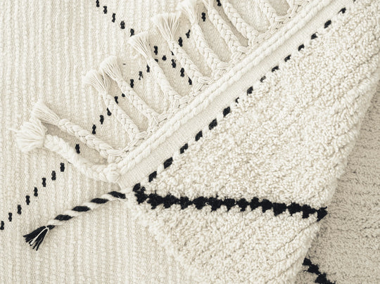 A cream wool berber rug with a black diamond pattern and plaited tassels folded over.