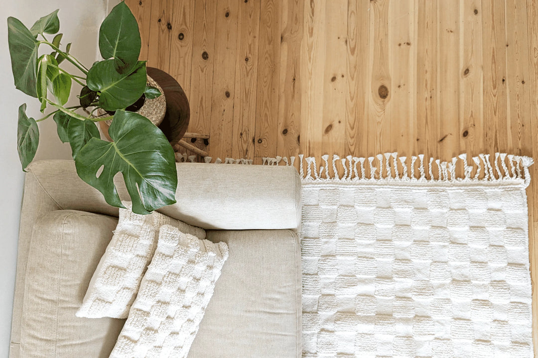 A checkered ivory wool berber rug with cotton plaited tassels on a wooden floor next to a sofa with matching wool cushions and a table with a plant on
