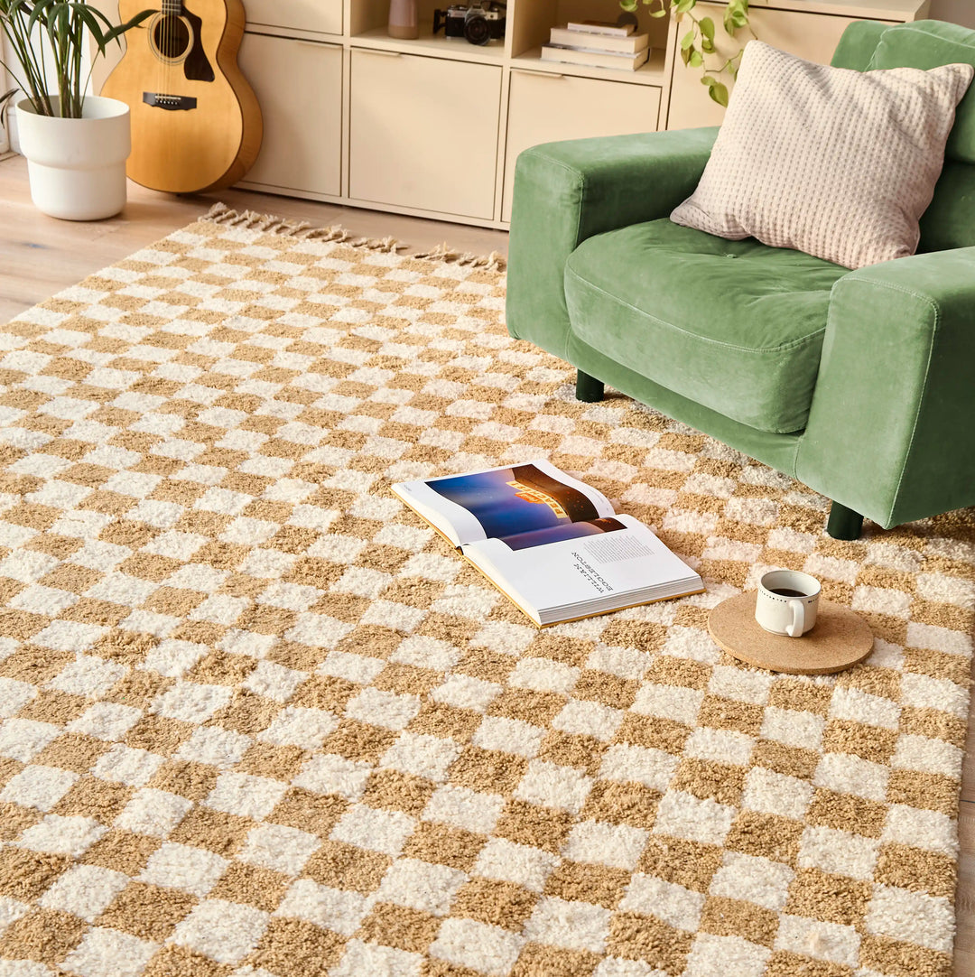 A checkered ochre berber rug with a green armchair, coffee cup and open book on top, in a sunlit room.