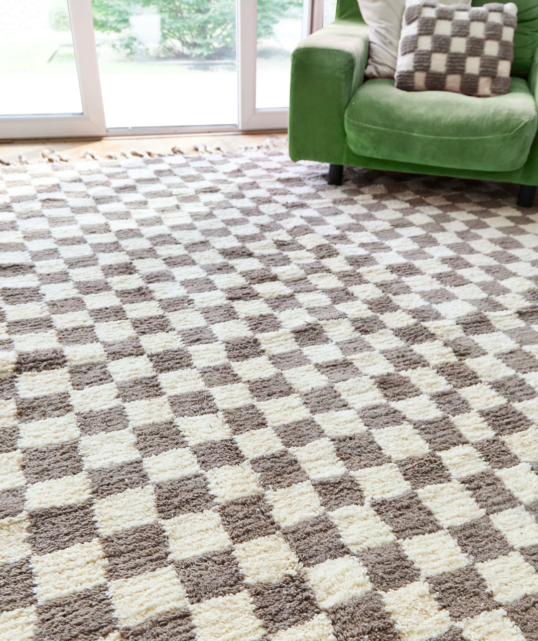 A checkered wool berber rug in taupe and cream on a wooden floor next to a green armchair and open patio doors