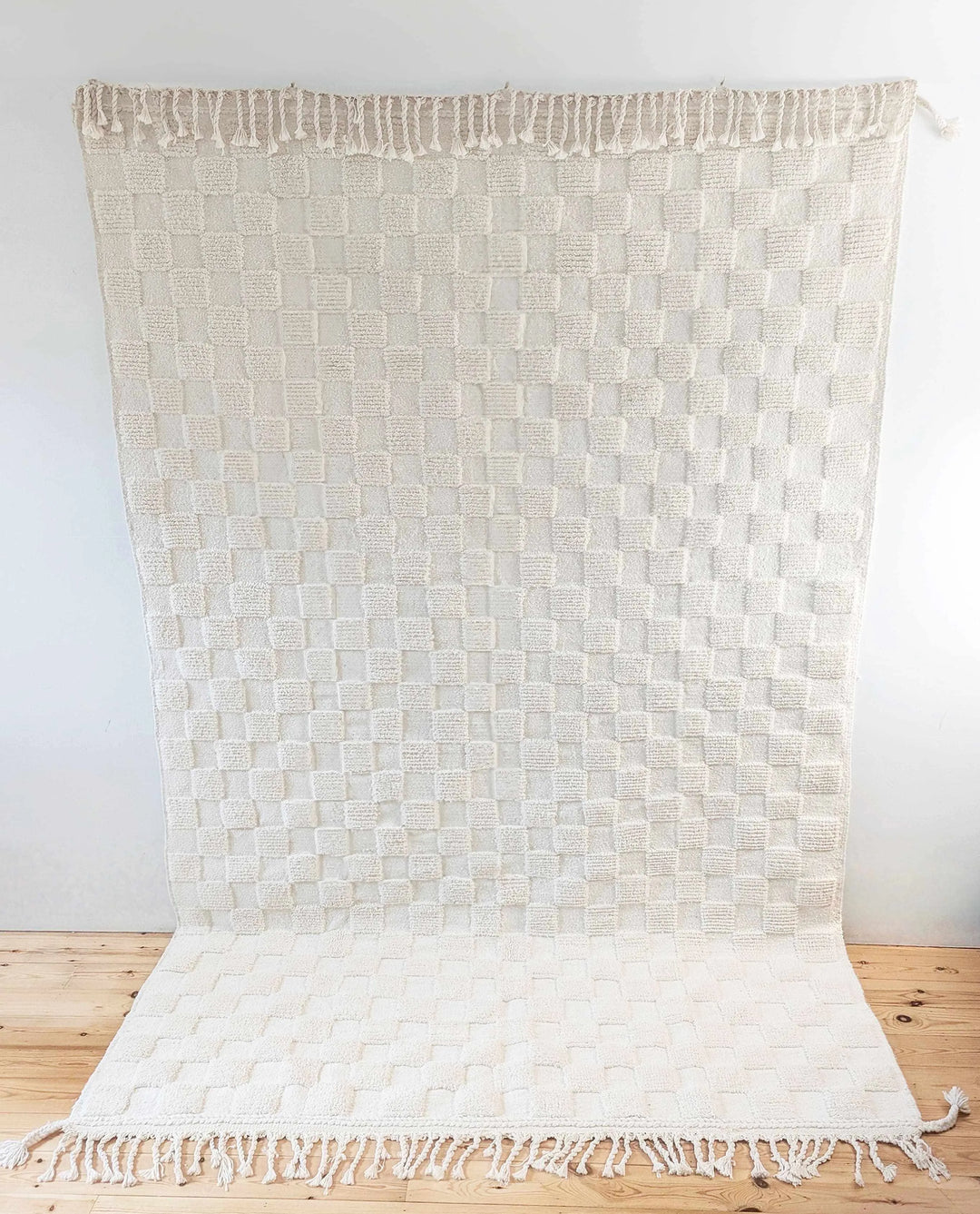 A checkered ivory wool berber rug with cotton plaited tassels hung up on a white wall above a wooden floor