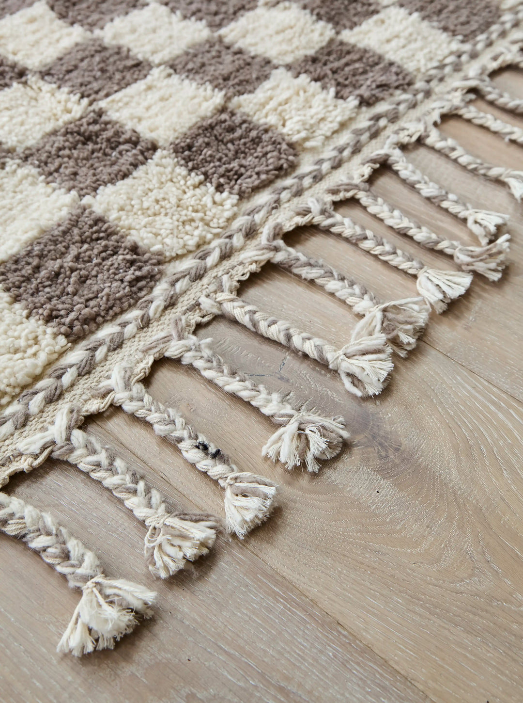 A checkered wool berber rug in taupe and cream on a wooden floor in close up on plaited cotton tassels