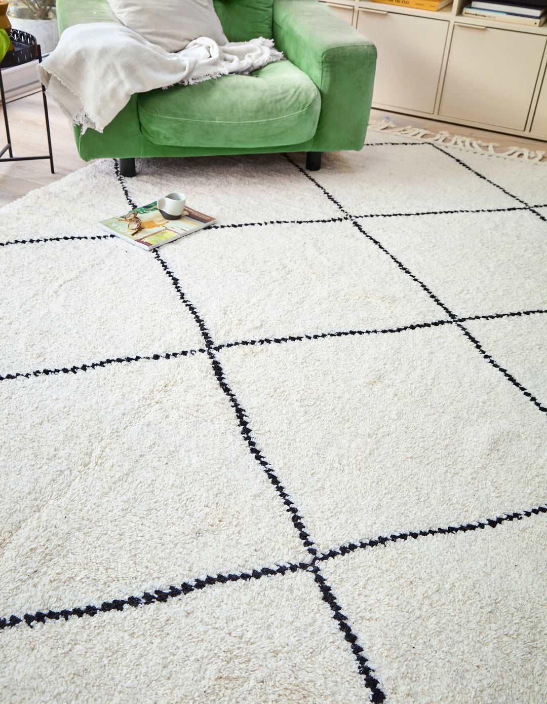 A cream wool berber rug with black diamond pattern in a sunlit room, with a green armchair, book and coffee on top 