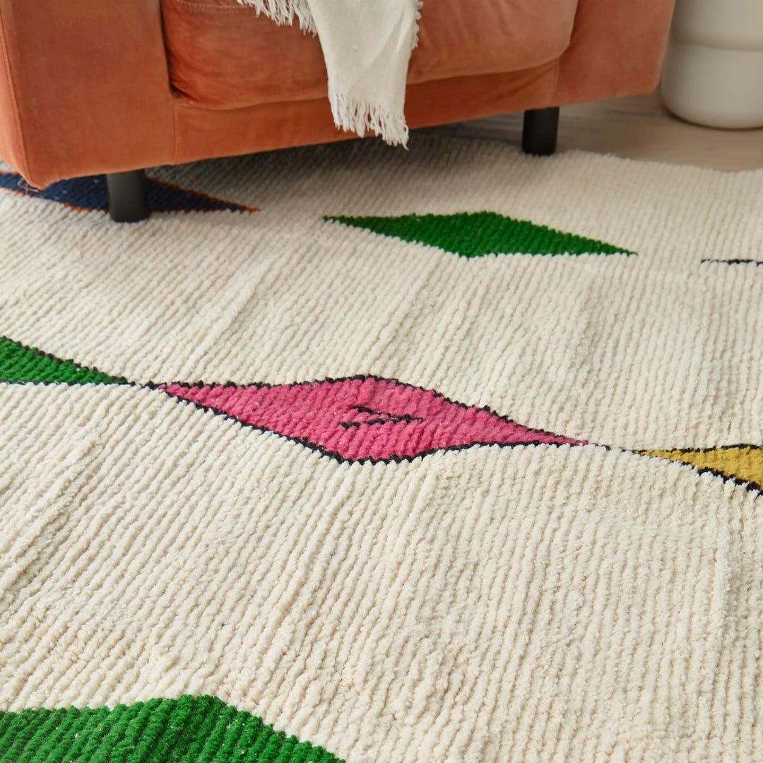 Close-up of a handwoven Berber rug featuring colourful diamond motifs, laid on wooden flooring near a pink armchair with a shawl on it