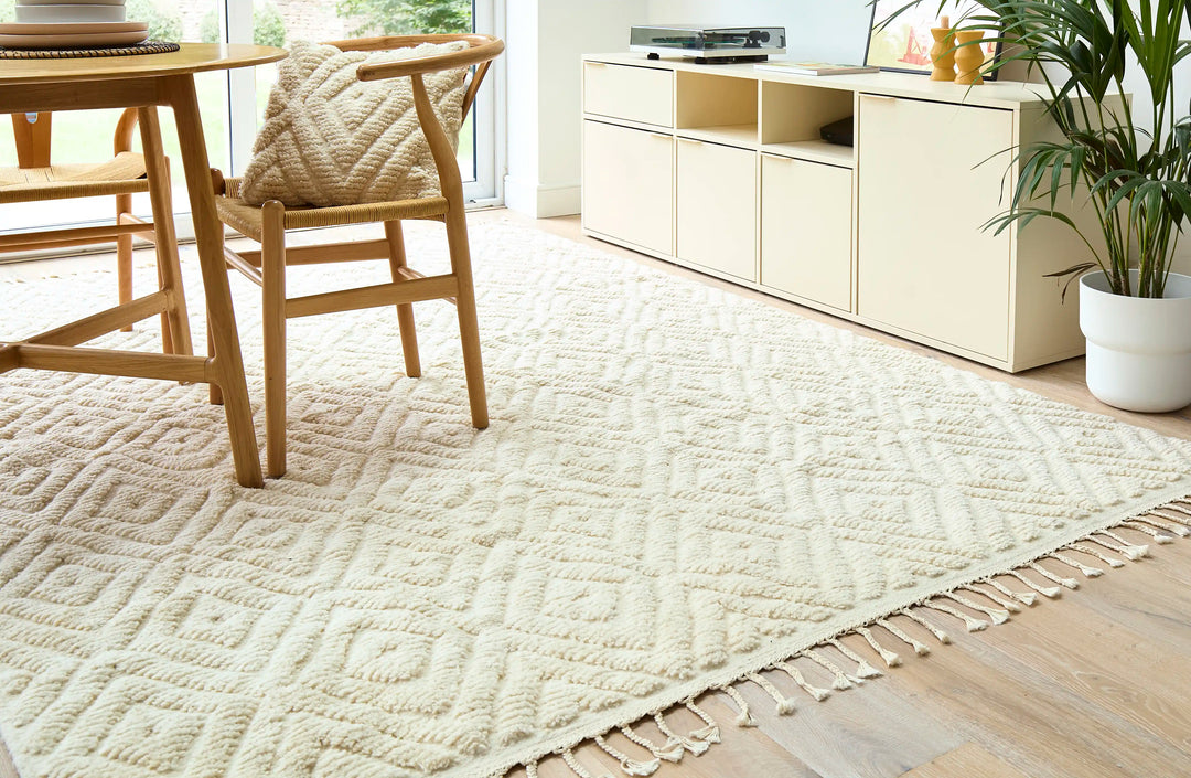 A natural wool cream berber rug with a diamond design on a wooden floor next to a patio door with a wooden table on top, next to cream cupboards and a plant.