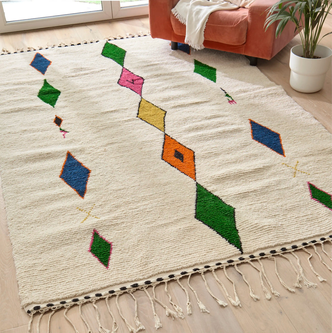 Handwoven Berber rug with vibrant multicoloured diamond pattern placed on wooden flooring next to a pink armchair and plant