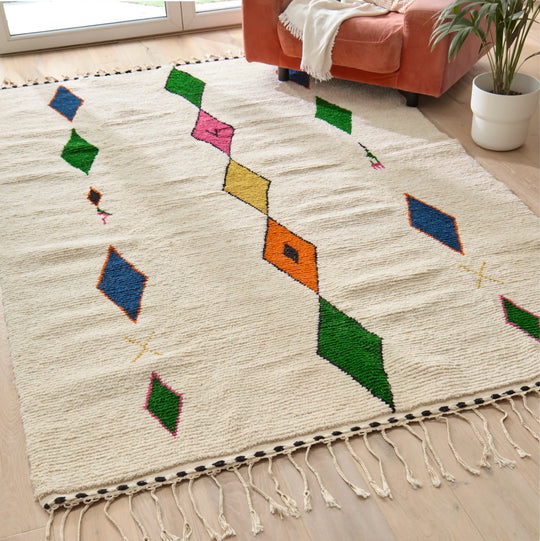 Handwoven Berber rug with vibrant multicoloured diamond pattern placed on wooden flooring next to a pink armchair and plant