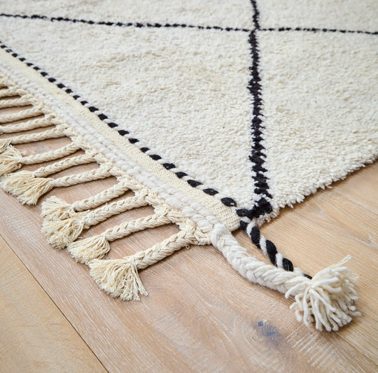 A cream wool berber rug with black diamond pattern and plaited tassels in close up on a wooden floor